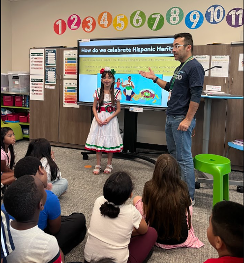 A teacher presents a lesson to a classroom of children. A student stands before a monitor displaying a presentation.