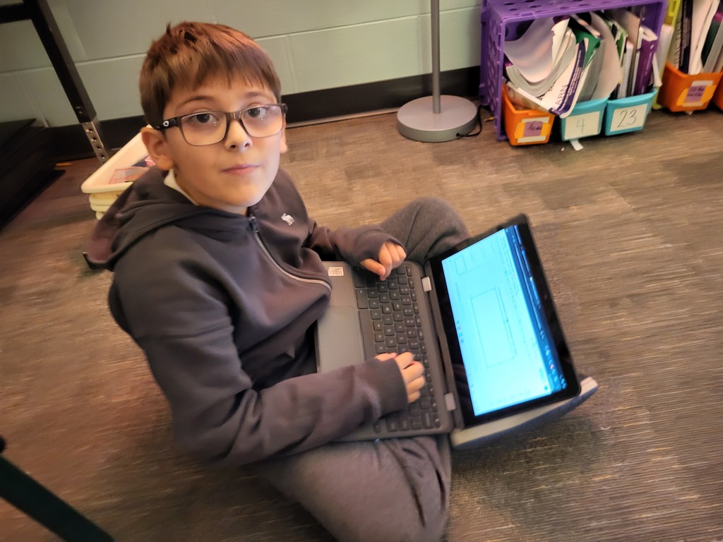 A student wearing glasses sits on the floor and uses a laptop with a screen showing text.