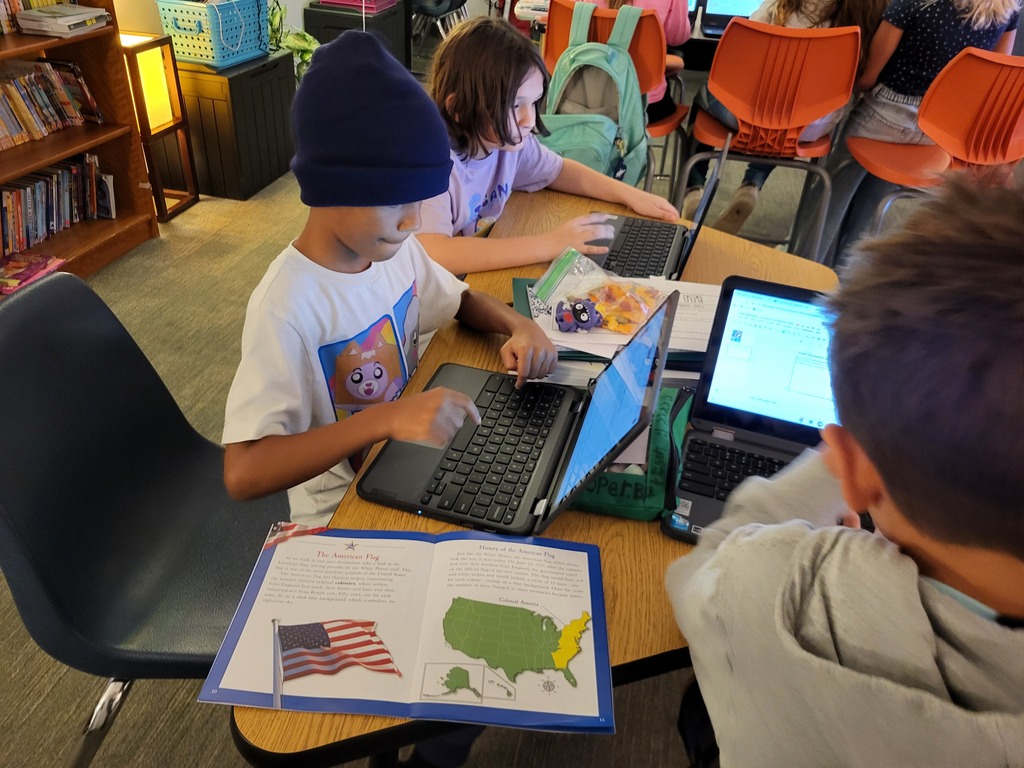 Four children using laptops in a classroom, one holding a book open to a map of the United States.