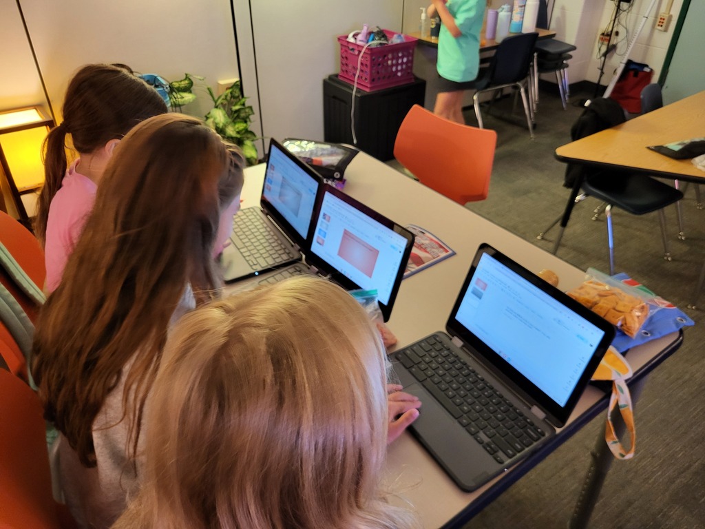 Three students seated at a desk with laptops and snacks. One person stands behind them.