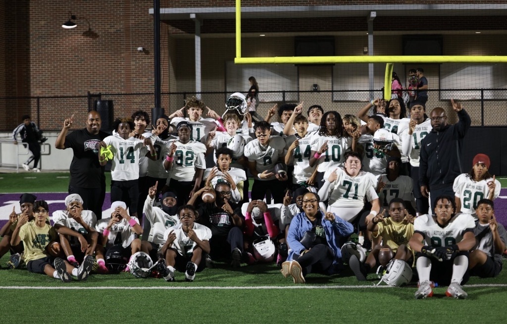 Football team posing on field, some in helmets, under yellow goalposts. Coach and adults stand nearby.