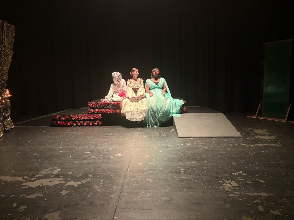 Three people sit on a bench on a stage. The person on the right wears a white mask.