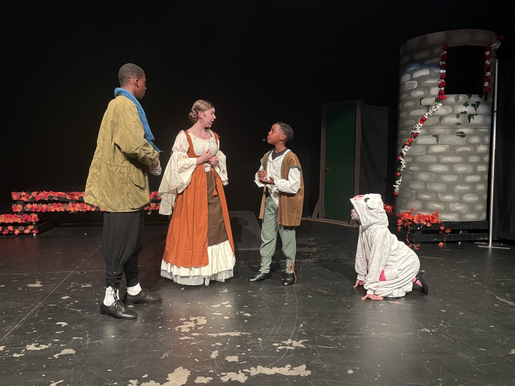 Four people in costumes on a stage with a backdrop. Three are standing, one kneeling, and there are two decorative structures behind them.