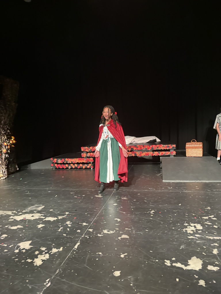Person in red cape and green skirt stands on stage with props and a wooden box nearby.