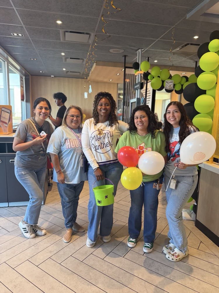 Five women stand near a counter with balloons. One holds a bucket, another holds a yellow balloon.