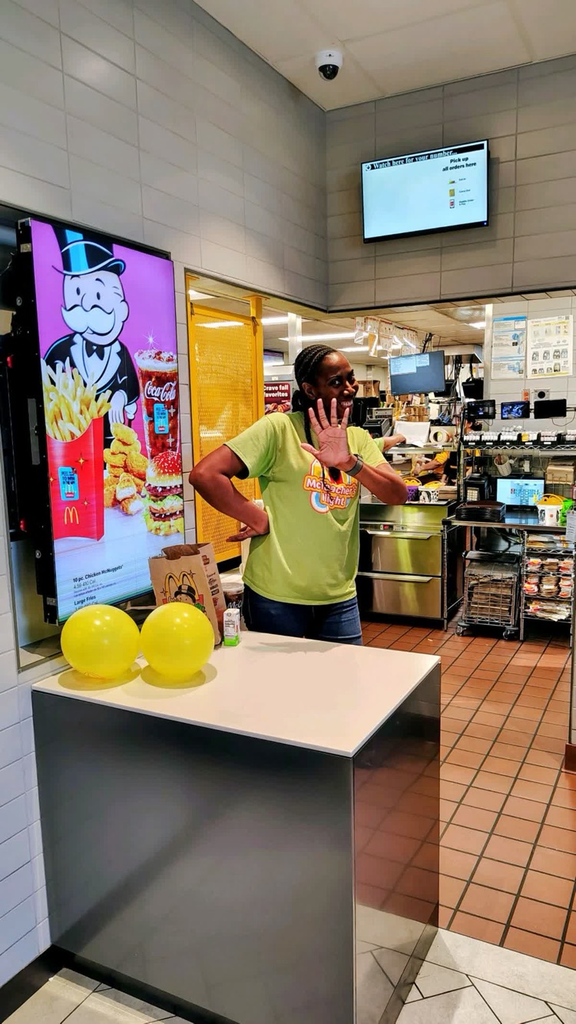 A woman stands at a counter inside a restaurant with a large screen showing a smiling man and fries behind her.