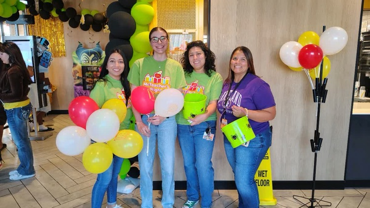 Five women in matching green shirts and blue jeans stand next to each other holding balloons and buckets.