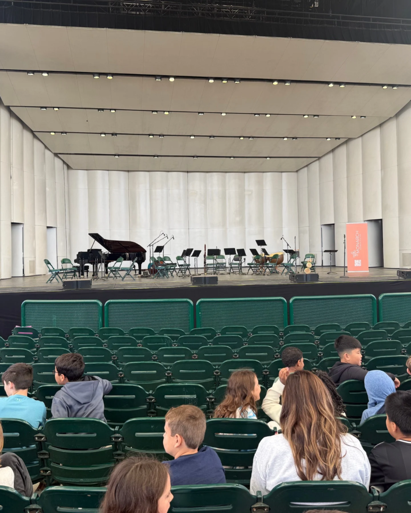 A large auditorium with a stage, a grand piano, and various musical instruments. Audience members are seated in green chairs.