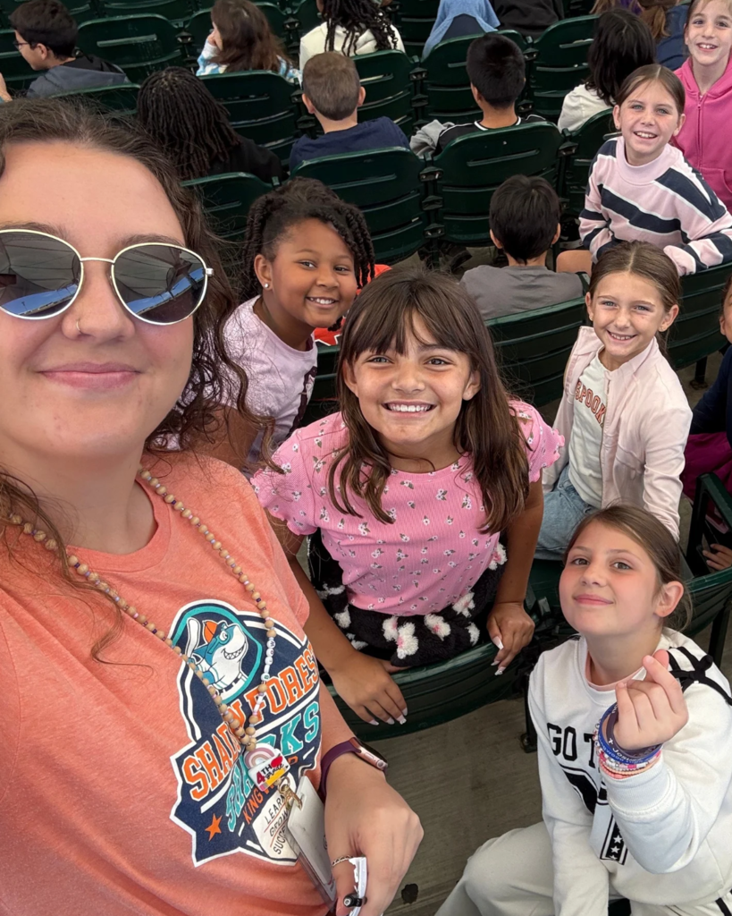 Woman and children in orange and white shirts sitting in stadium seats, smiling and posing for a photo.