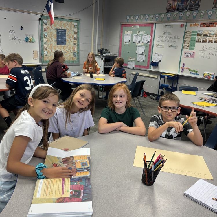 Four children sitting at a table in a classroom, smiling, with papers, pencils, and notebooks.