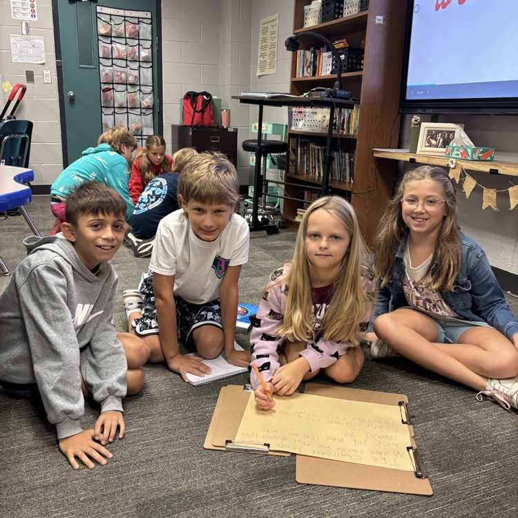 Four children seated on the floor in a classroom. Each holds a paper. Behind them, a monitor displays text.