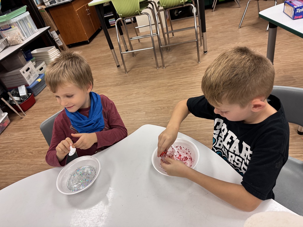 Two young boys sitting at a table, each with a bowl, one with red sprinkles, mixing contents.