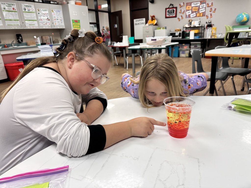 Two girls in a classroom are looking at a beaker of liquid on a table.