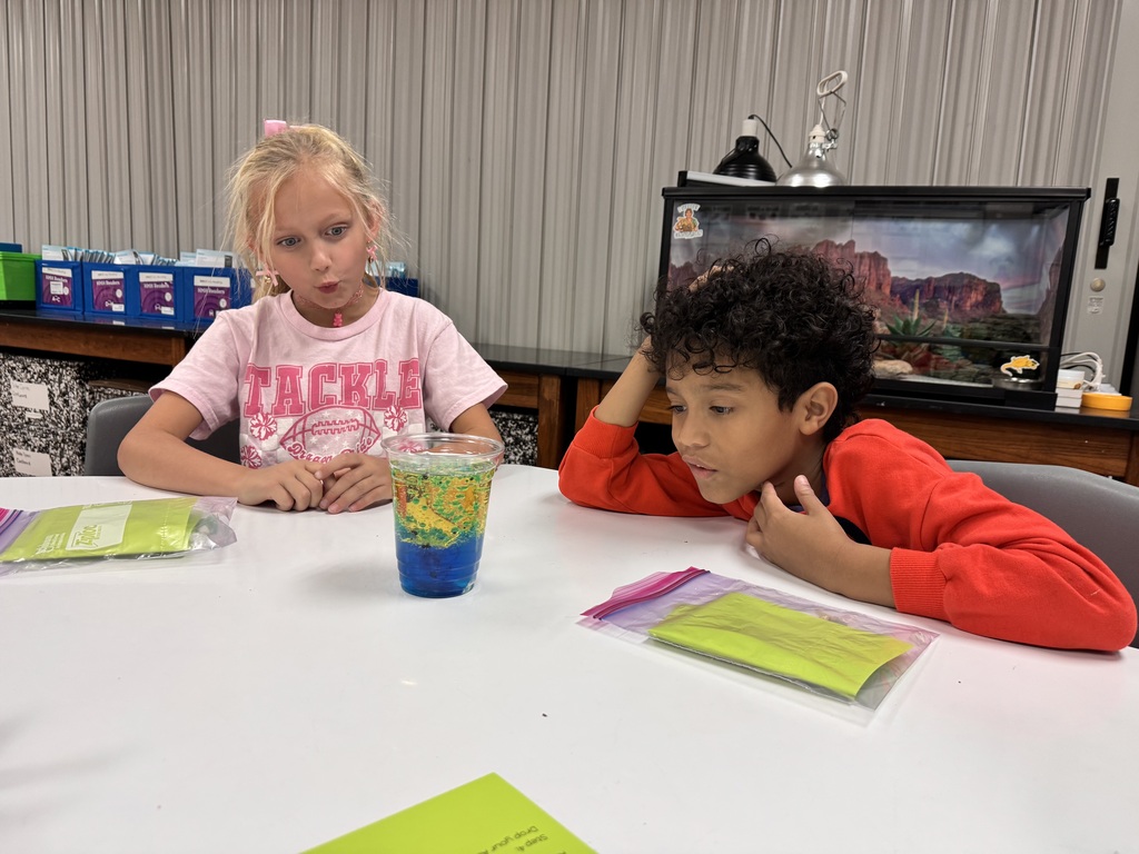 Two children seated at a table, facing a blue liquid in a clear glass. One child holds a plastic bag.