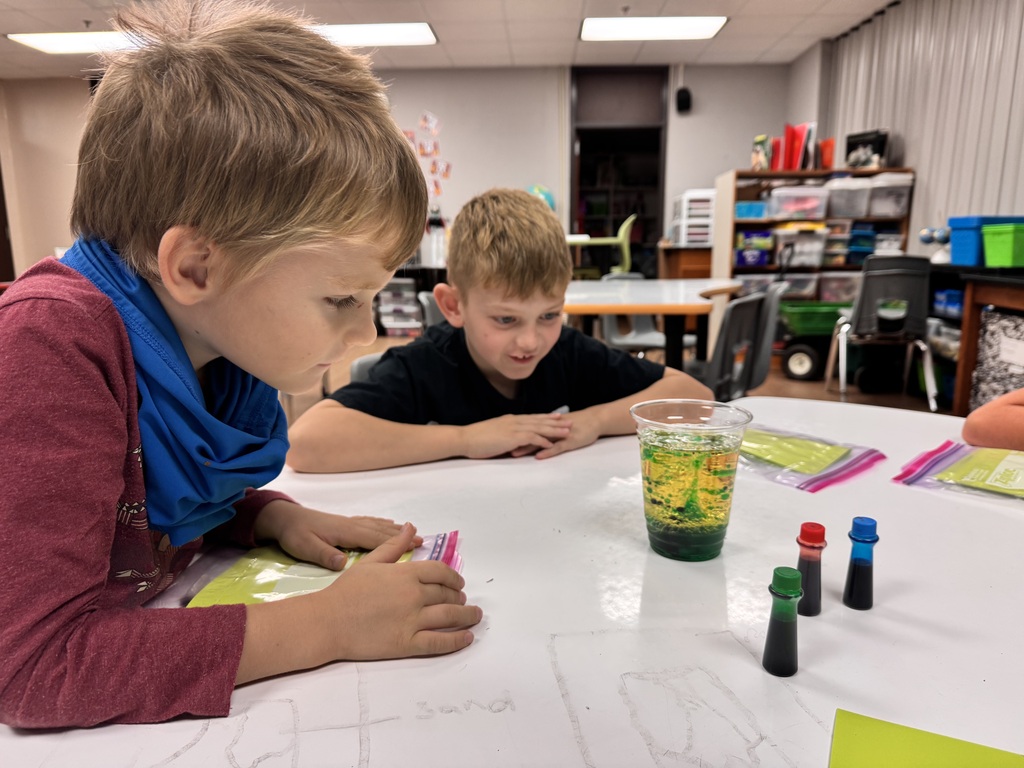 Two boys work on a table with a cup of liquid, markers, and a yellow paper.