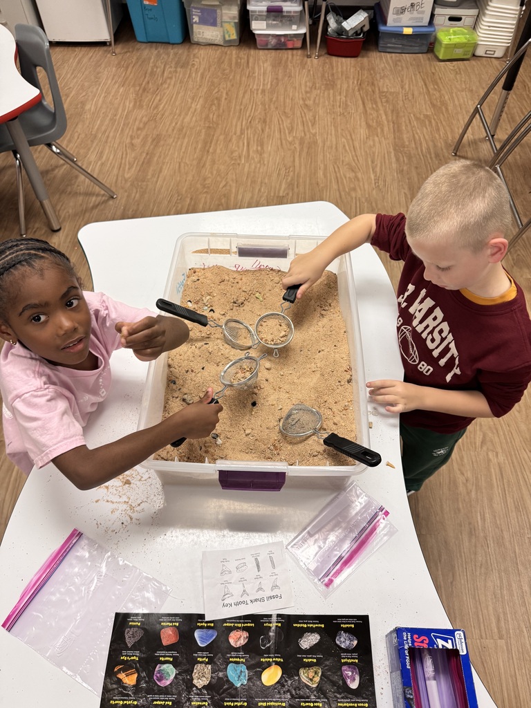 Two children play with sand and metal tools in a large container on a white table.