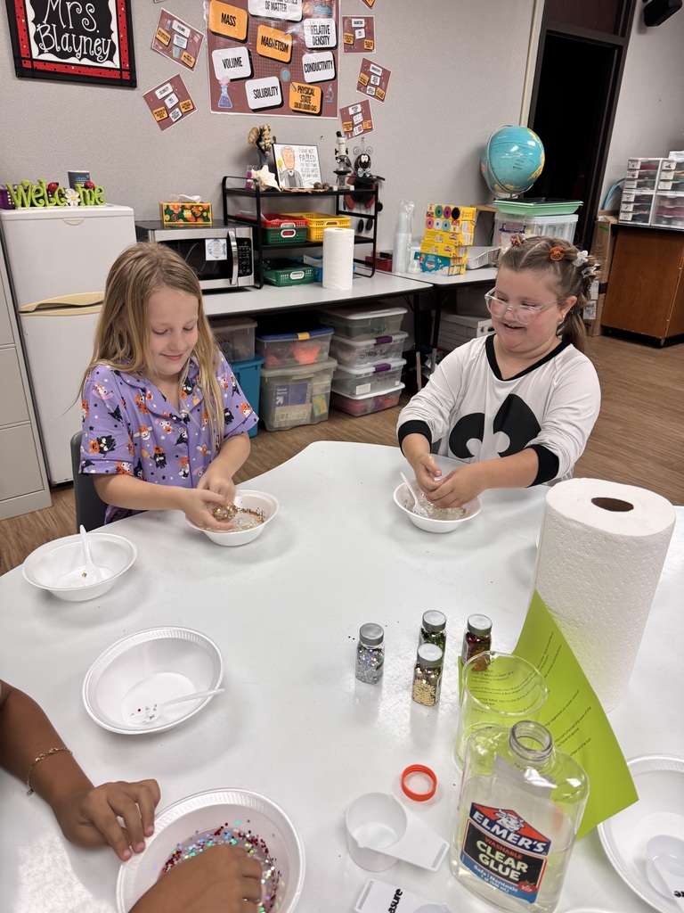 Two girls sitting at a table with bowls, bottles, and a paper towel roll. One girl is wearing glasses.