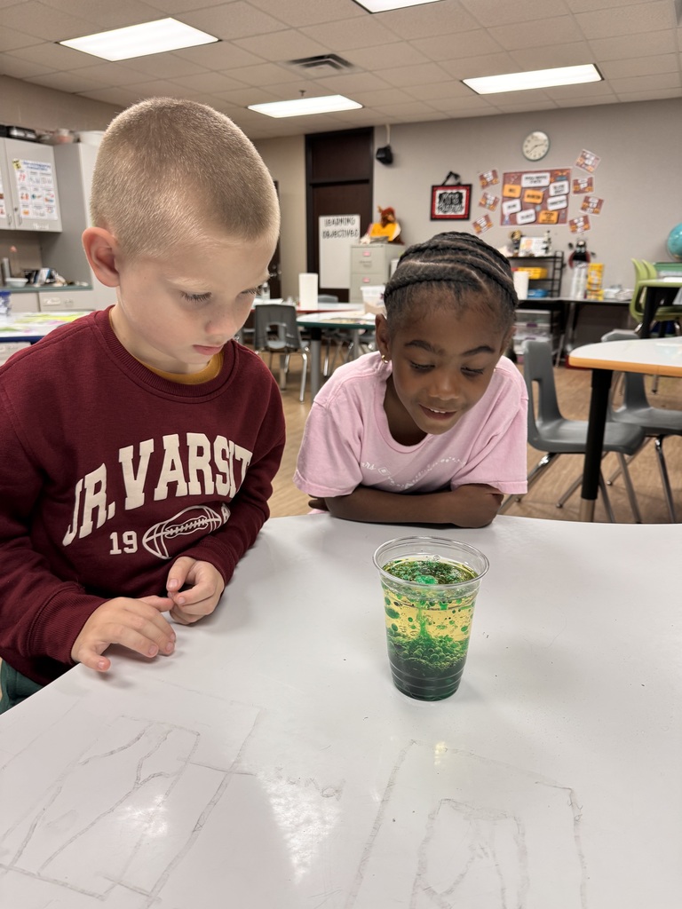 Two young children are working together at a table. One has a cup with layered colored liquids.