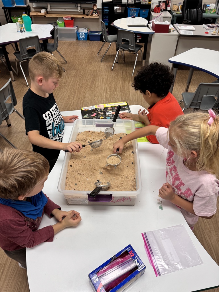 Four children seated at a table, playing with sand in a large container with various tools.