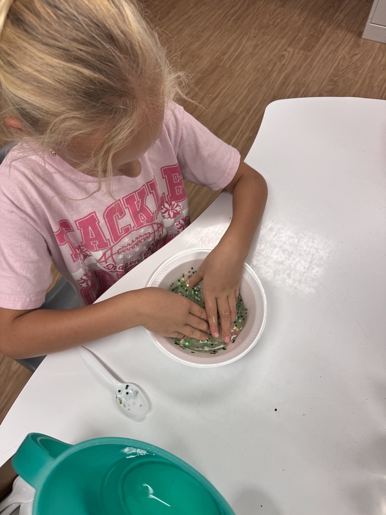 A girl sits at a table, mixing green substance in a bowl with her hands.