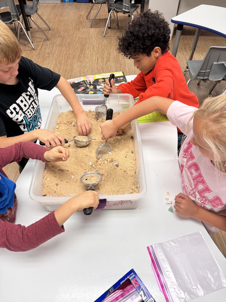 Four kids at a table, sifting sand through a small bucket using a small shovel.