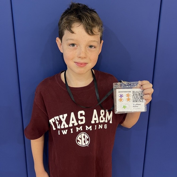 Boy in maroon shirt holding a badge, standing against blue wall, smiling, with a lanyard around his neck.
