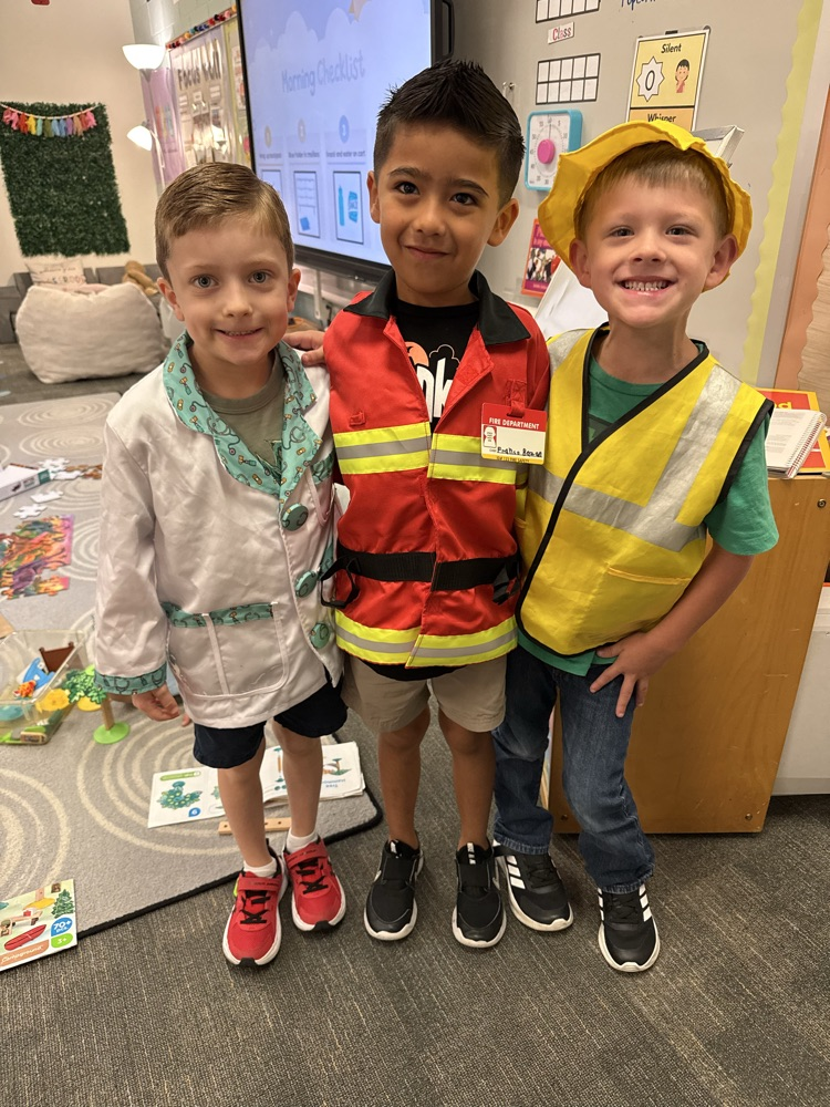 Three young boys wearing safety vests stand together indoors, smiling, in front of a large screen.