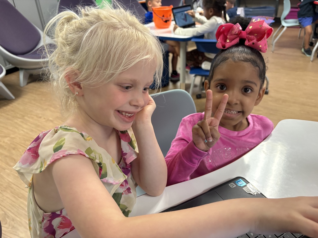 Two young girls sit at a table, one wearing a floral dress, the other in pink. They face a laptop.