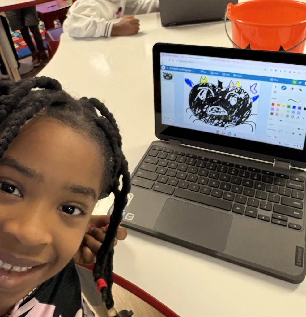 A child sits at a desk with a laptop displaying a cartoon face. A bucket is on the desk.
