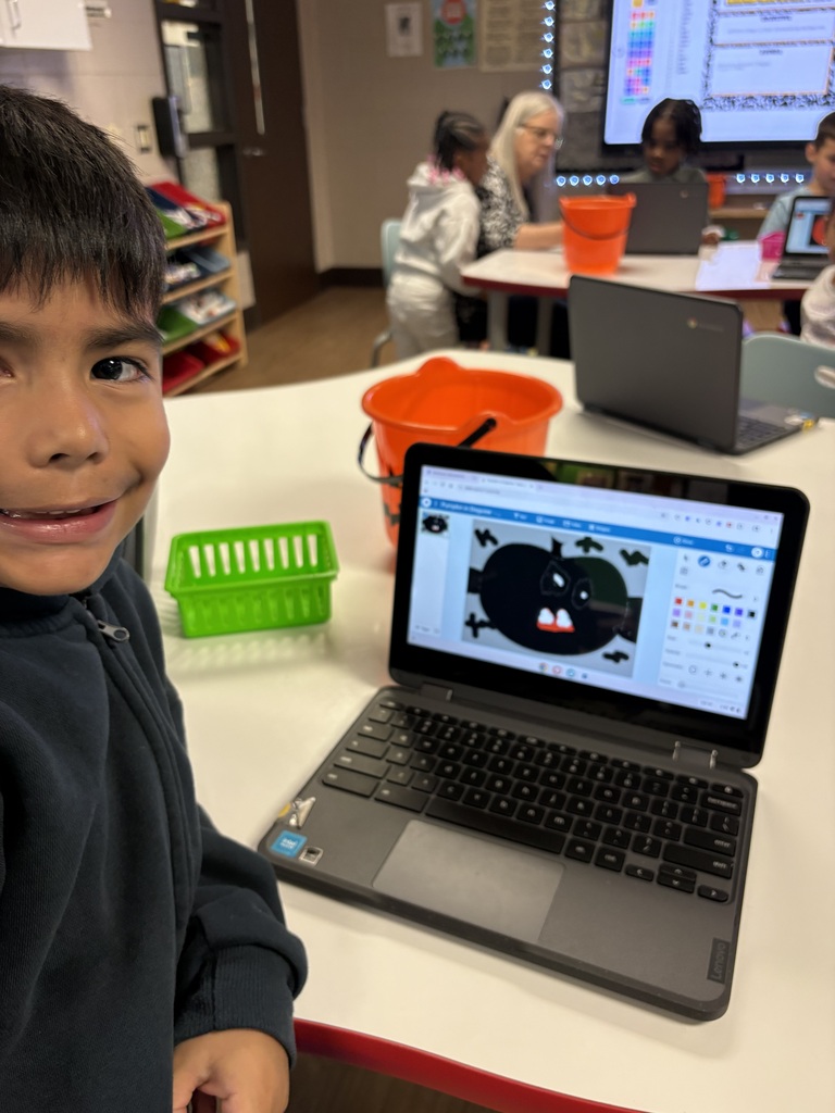 Child smiling, standing next to a laptop, with a green basket, bucket, and other desks.
