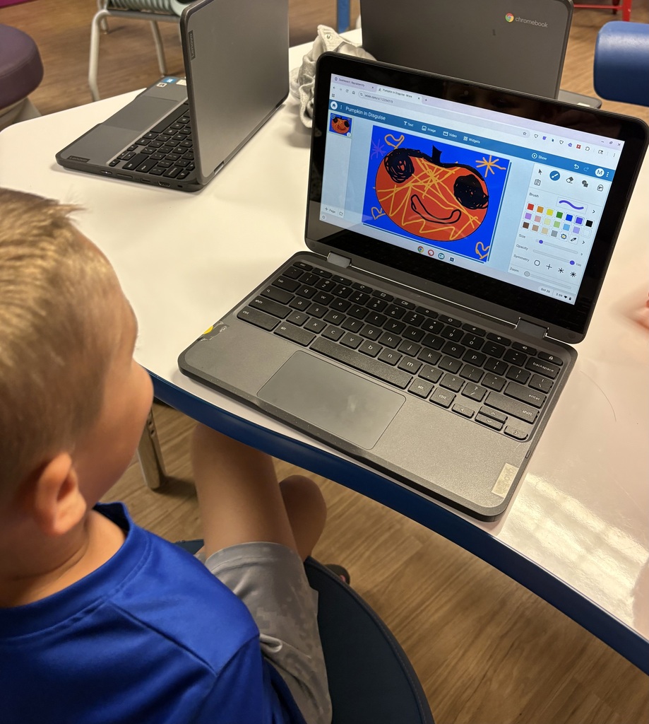 A child seated on a chair at a desk with laptops. One laptop shows a Halloween pumpkin.