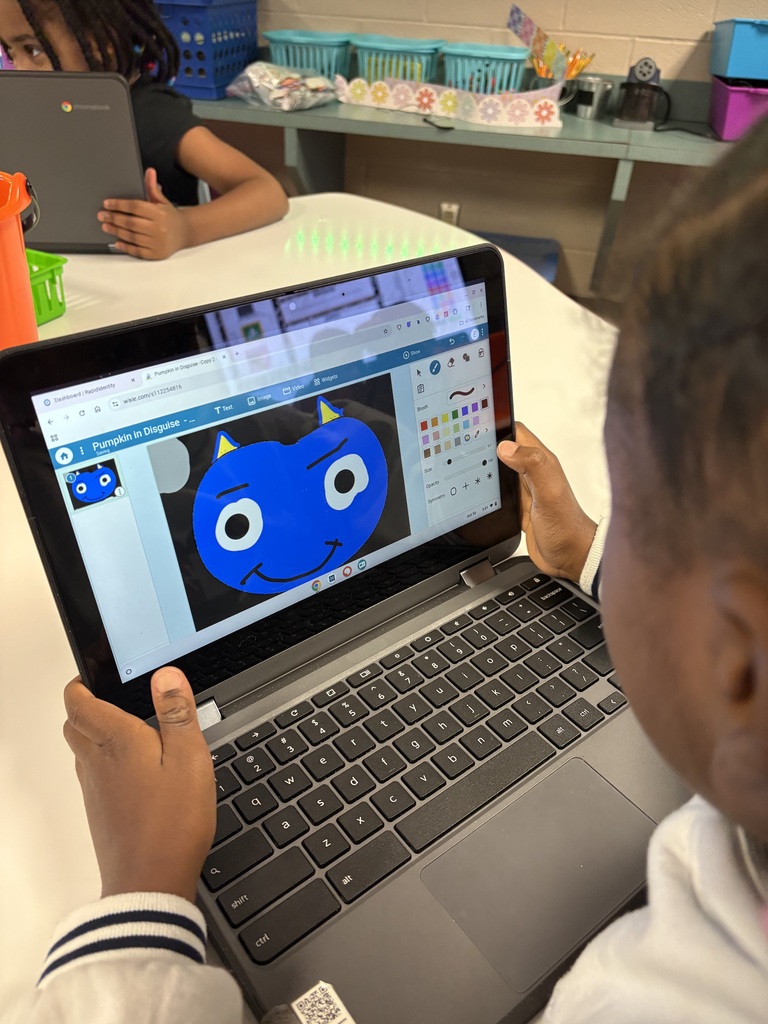 A student sits at a table using a laptop with a blue cartoon character on screen.