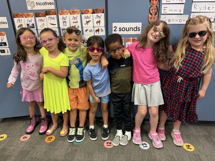 Seven children stand in a row, wearing sunglasses, in a classroom. Behind them is a wall with name labels.