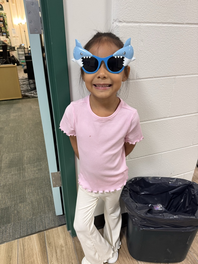 Young girl in pink shirt and sunglasses with shark ears standing near a door.