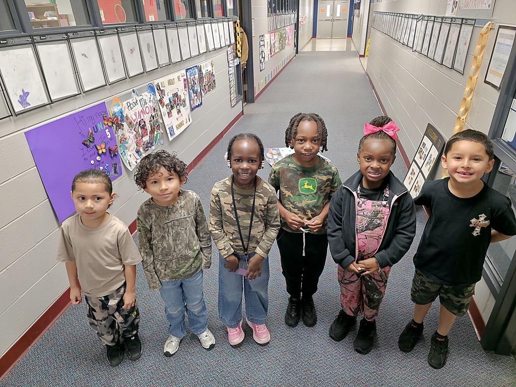 Six children standing in a hallway with colorful wall art. They wear various shirts and shoes, with one holding a camera.