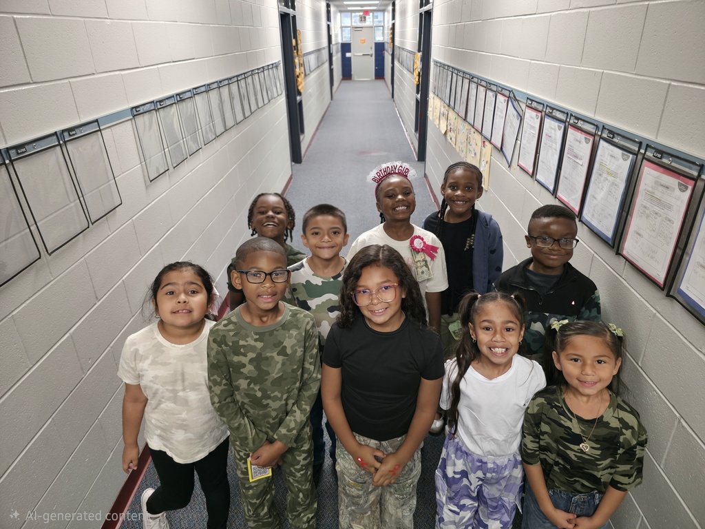 Group of students in a hallway, smiling at the camera, some wearing glasses. White walls and papers.