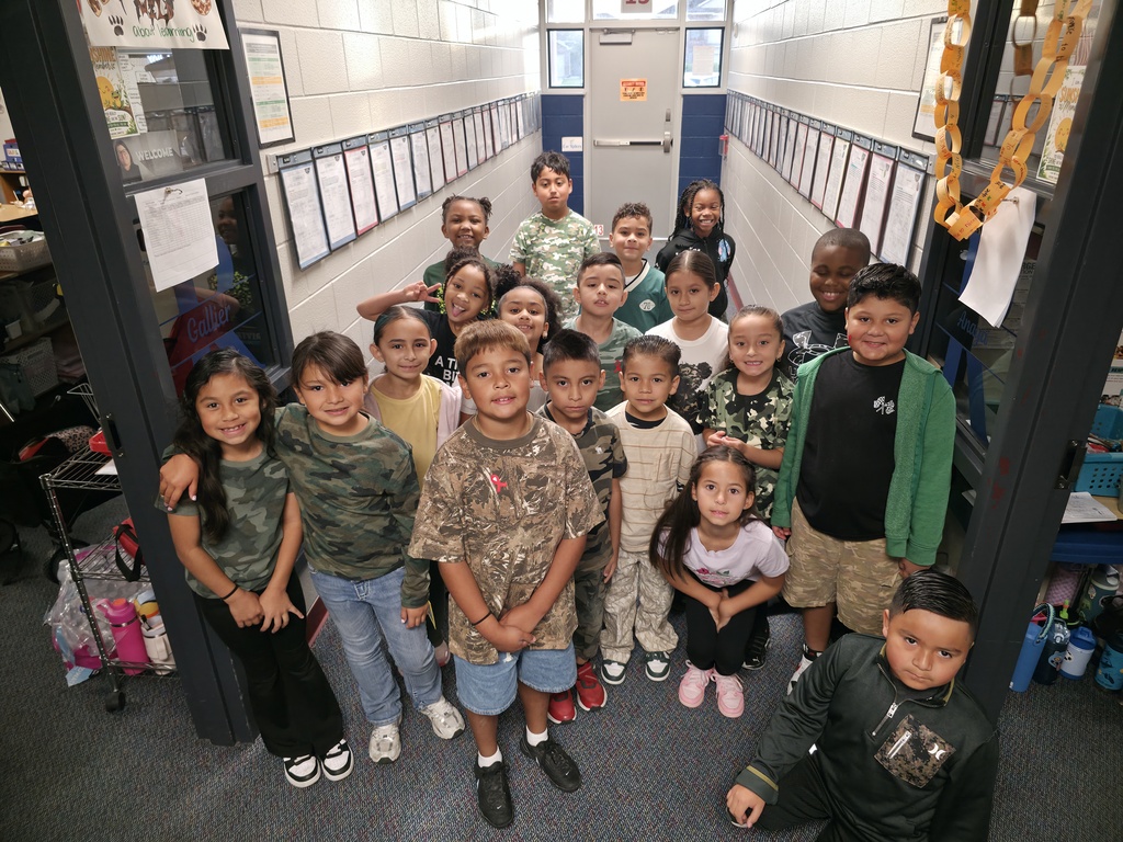 A group of children stands in a hallway with bulletin boards on both sides. Some have backpacks.