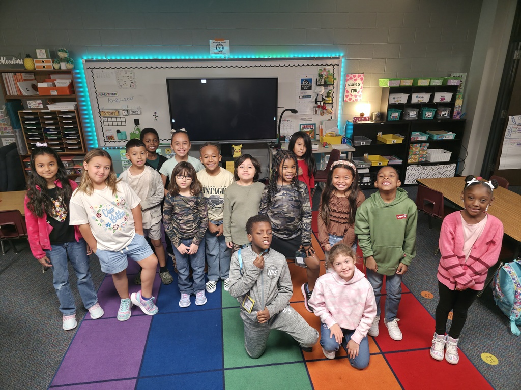 Students in casual clothing stand and crouch on a colorful rug in a classroom. Behind them is a television and shelves with school supplies.
