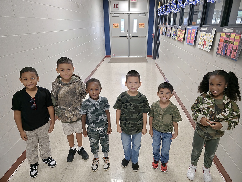 Six children in matching camouflage clothing stand in a school hallway, smiling at the camera.