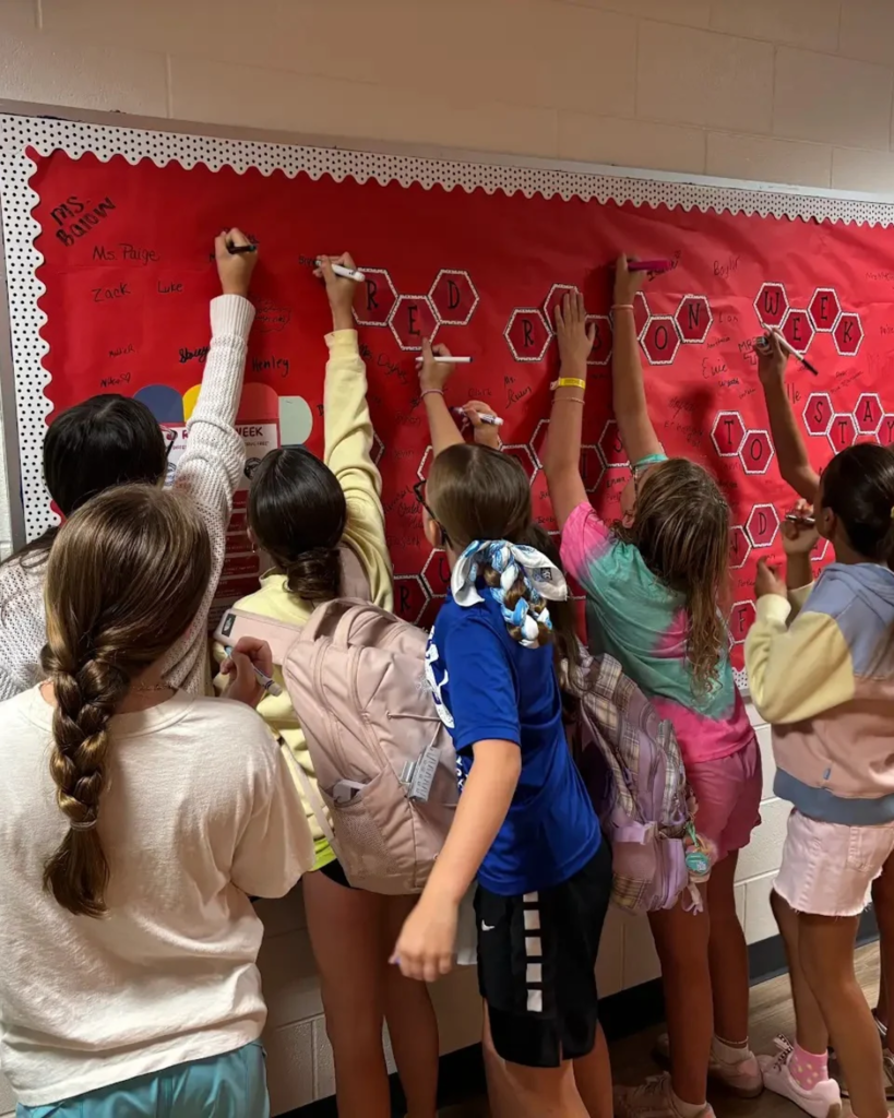 Students stand at a bulletin board, each holding a marker, writing on a red poster with hexagons.