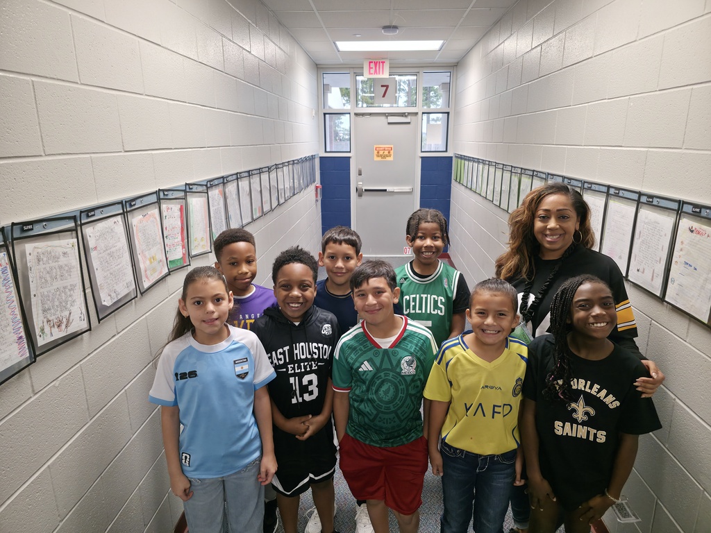 Group of children wearing sports jerseys pose in a brightly lit corridor with white walls and bulletin boards.