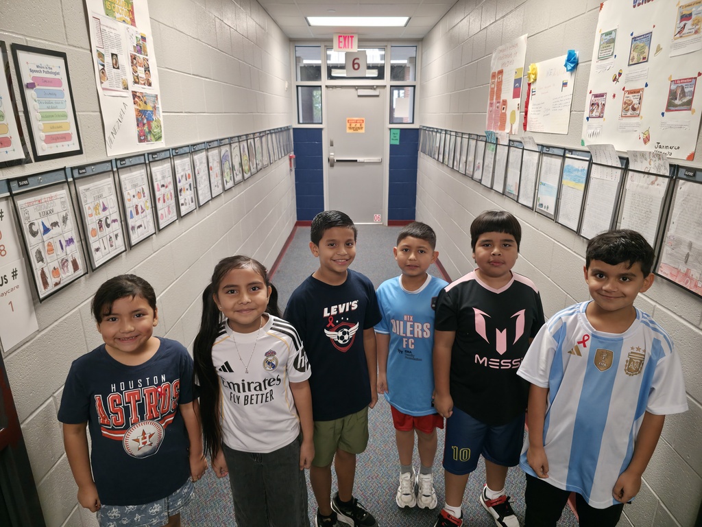 Five children stand in a hallway. They wear various colored shirts. The hallway has bulletin boards with posters and papers.