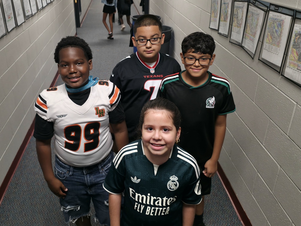 Five children in sports uniforms standing together in a hallway with picture frames on the walls.