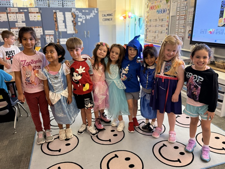 A group of children dressed in various costumes stand in a classroom. Some wear shoes, others slippers.