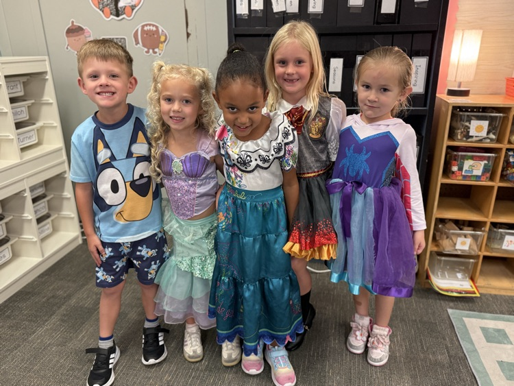 Five children wearing costumes, two girls in dresses and three boys in casual clothes, standing in a classroom with shelves and a lamp.