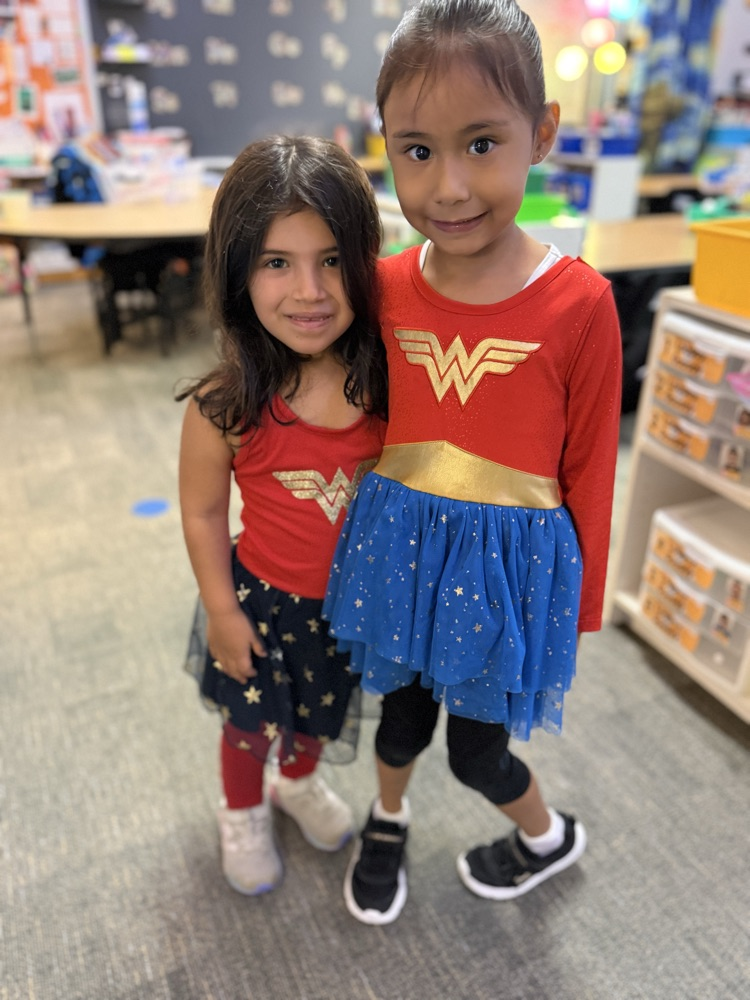 Two young girls wearing matching Wonder Woman shirts stand next to each other in a classroom.