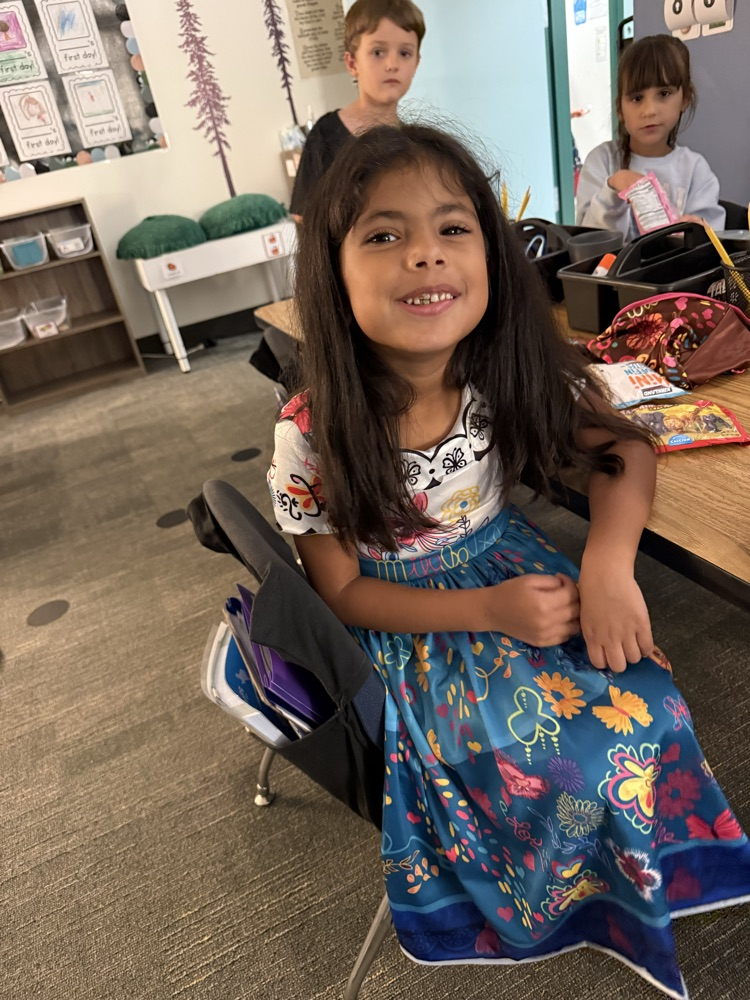 A girl in a floral dress sits in a chair at a classroom table with another child.
