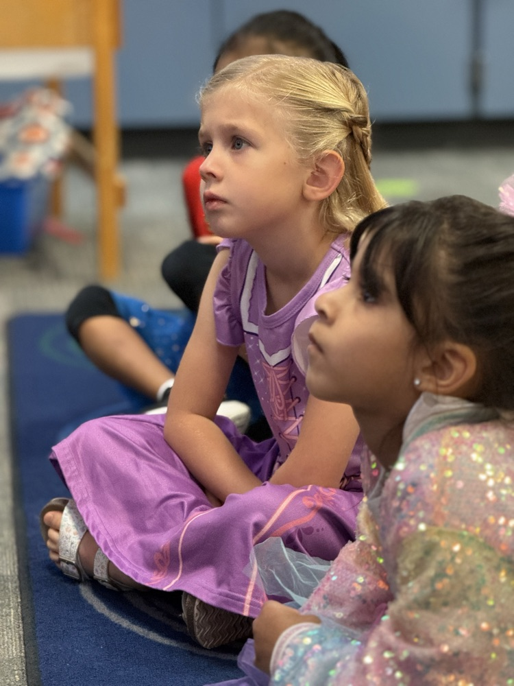 Students sit on the floor in a classroom, one wearing a purple dress and the other in a sequined outfit.