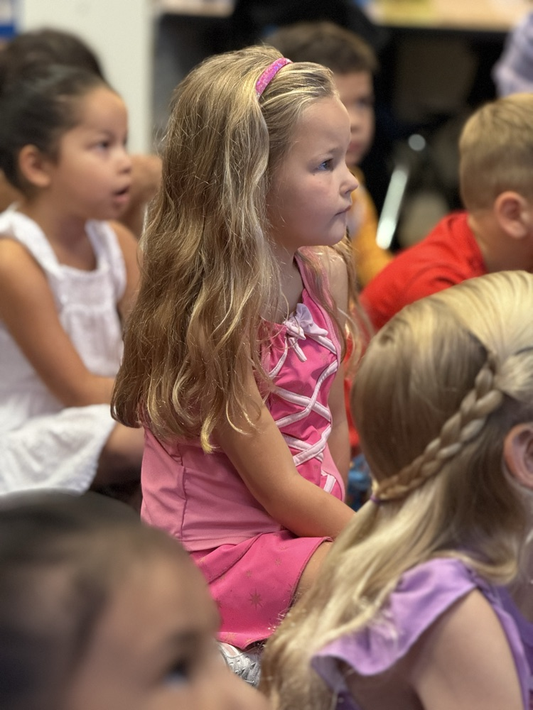 Several children sit attentively in a classroom, with one child in pink and another in white.