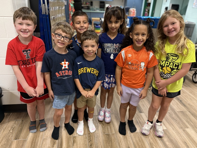 Nine children stand together on a wood floor. Some are wearing glasses. The children are smiling.